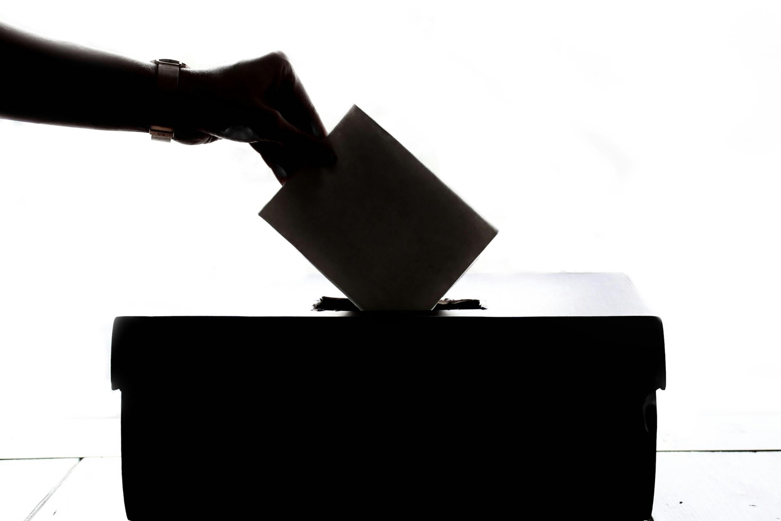 photo in shadow of hand placing a ballot into a ballot box, symbolizing the right to vote and a democracy that allows people to safely and freely cast their vote for the candidates of their choosing